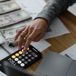 Pessoa usando a calculadora do celular ao lado de dinheiro e documentos financeiros sobre uma mesa.