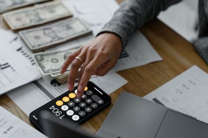 Pessoa usando a calculadora do celular ao lado de dinheiro e documentos financeiros sobre uma mesa.