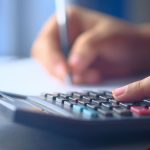 Mãos fazendo cálculos em uma calculadora e anotando informações para planejar o orçamento doméstico.