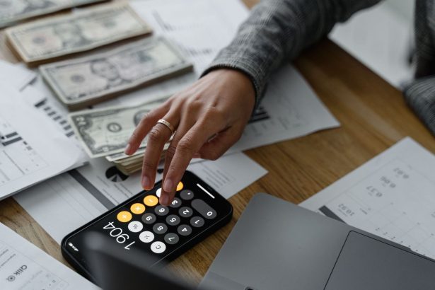 Pessoa usando a calculadora do celular ao lado de dinheiro e documentos financeiros sobre uma mesa.