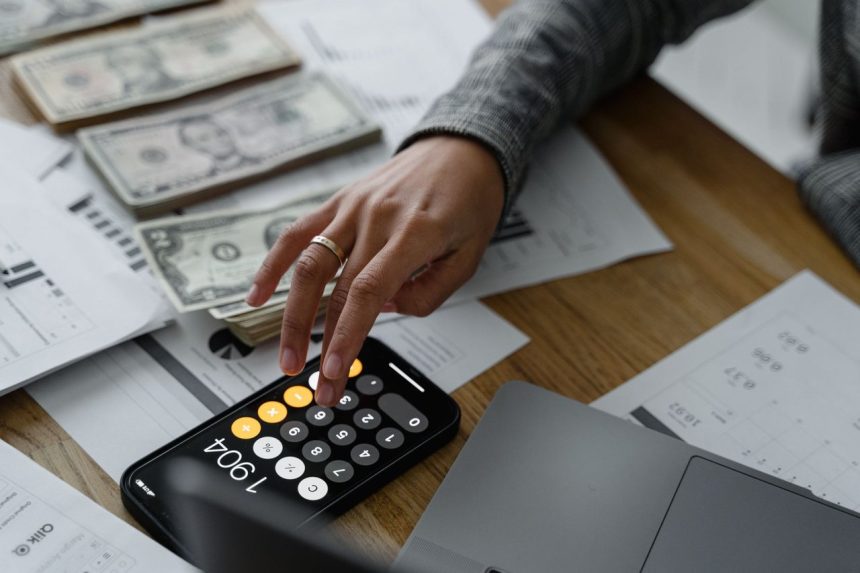 Pessoa usando a calculadora do celular ao lado de dinheiro e documentos financeiros sobre uma mesa.