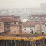 Trabalhadores atuam em um canteiro de obras urbano, com edifícios ao fundo.