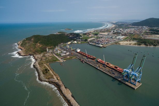 Vista aérea do Porto de Imbituba, com cais, guindastes, embarcações e a linha costeira ao redor.