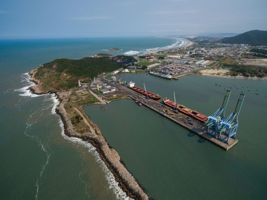 Vista aérea do Porto de Imbituba, com cais, guindastes, embarcações e a linha costeira ao redor.