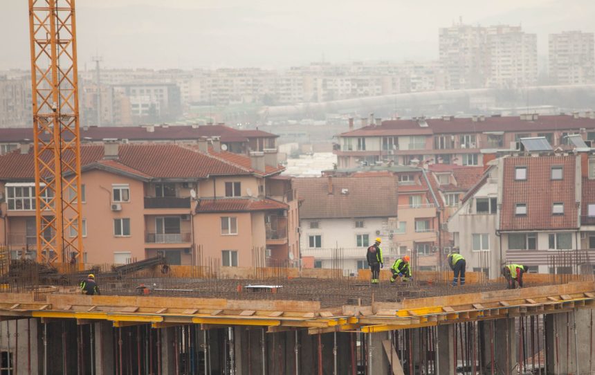 Trabalhadores atuam em um canteiro de obras urbano, com edifícios ao fundo.